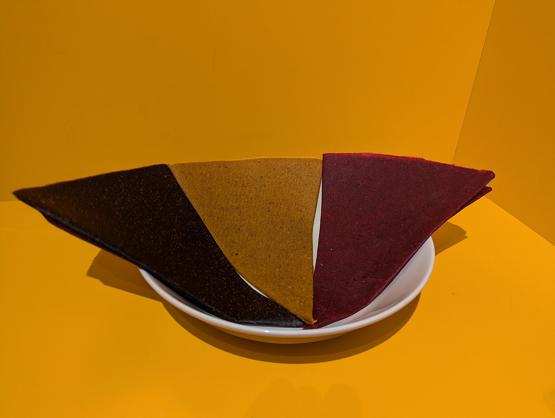 Three triangles of tklapi fruit leather in dark plum, golden and red flavors in a white bowl, bought at Dezerter Bazaar in Tbilisi