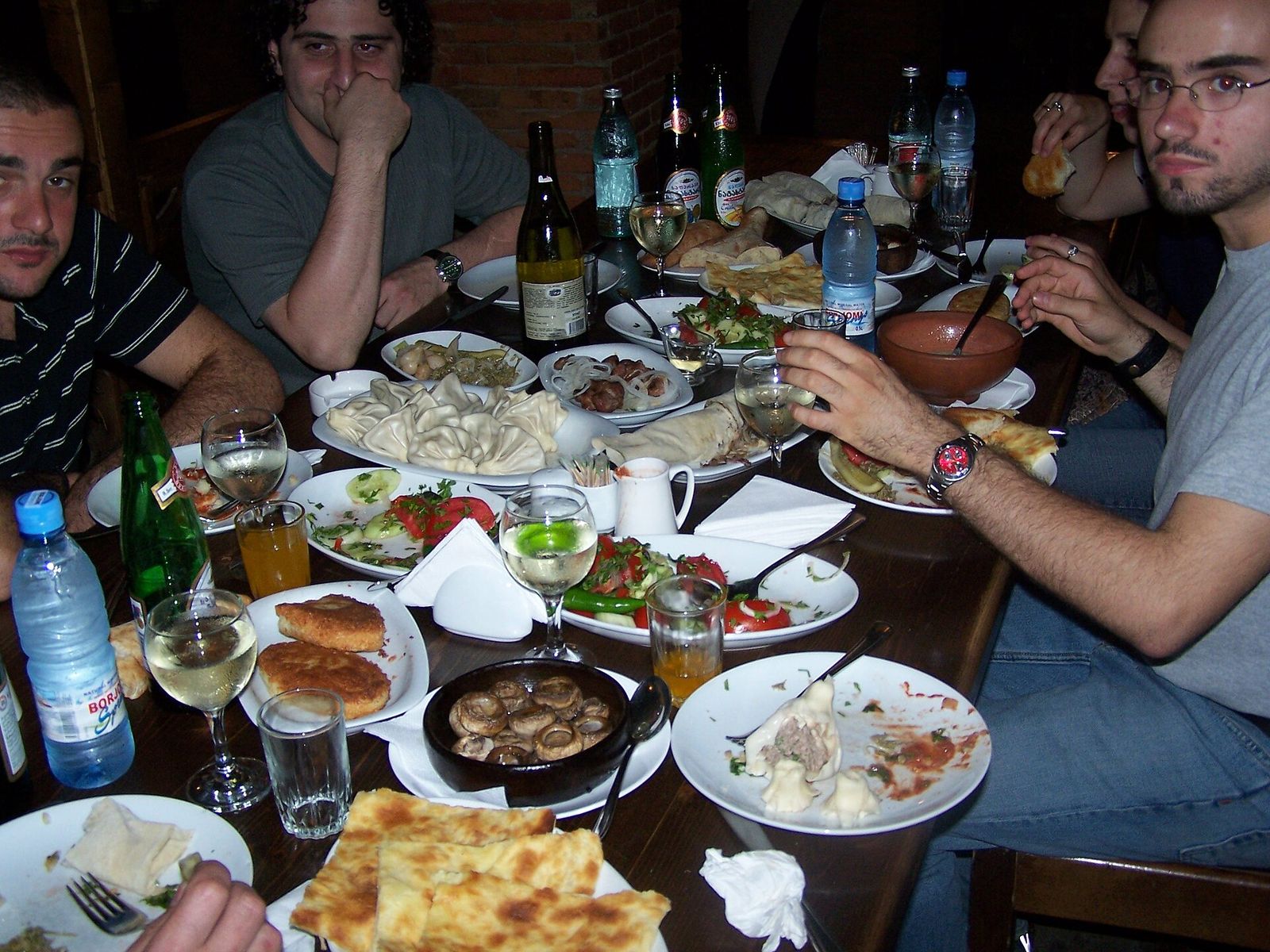 A Georgian supra table crowded with plates of khinkali, khachapuri, salads, mushrooms in a clay dish, wine glasses and bottles