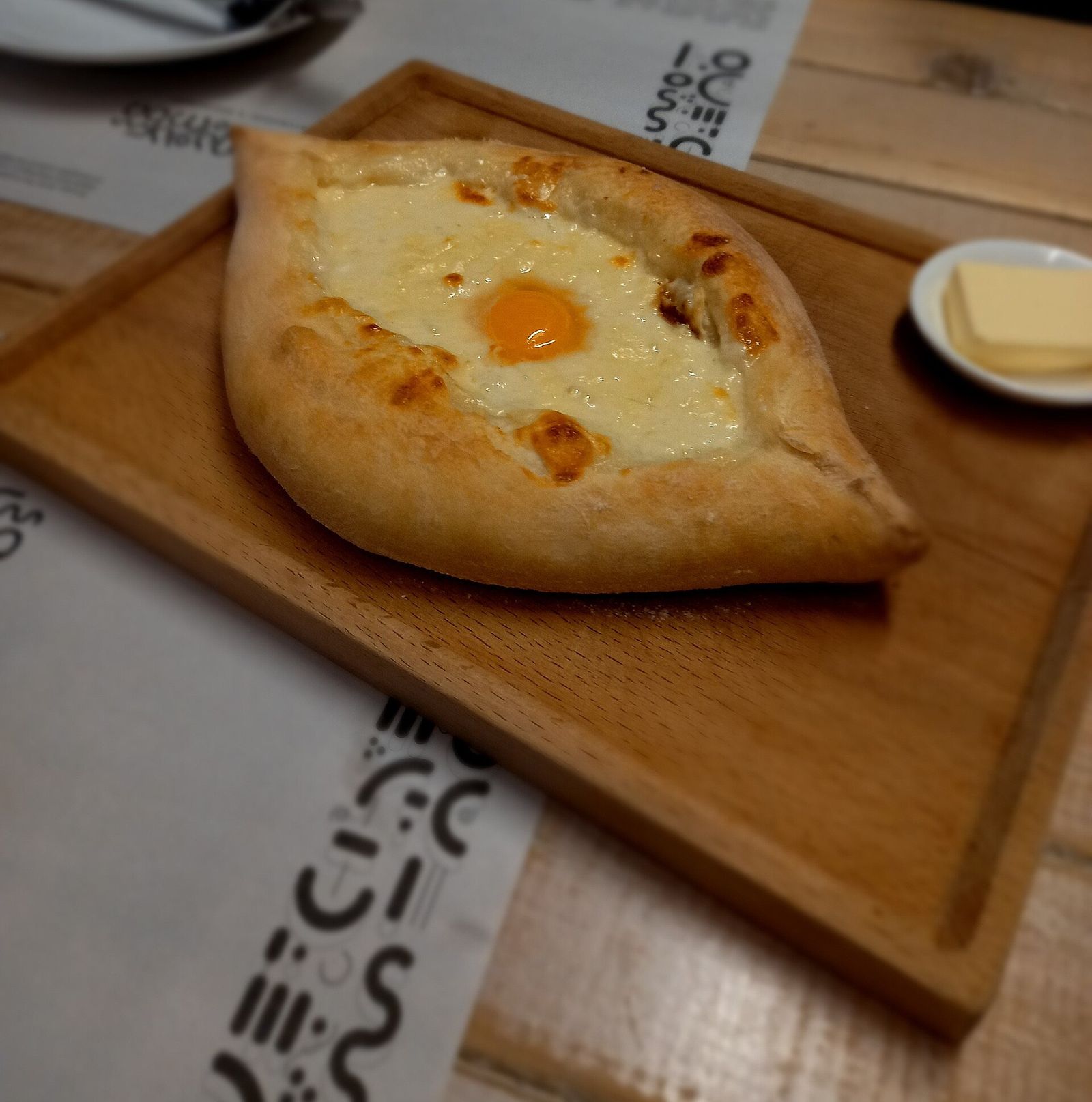 Adjaruli khachapuri, the boat-shaped cheese bread with an egg yolk in the middle, on a wooden board with a dish of butter