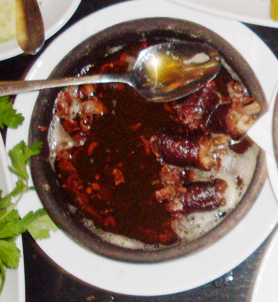 Pieces of kupati sausage fried in a clay dish with their juices, a spoon resting on top