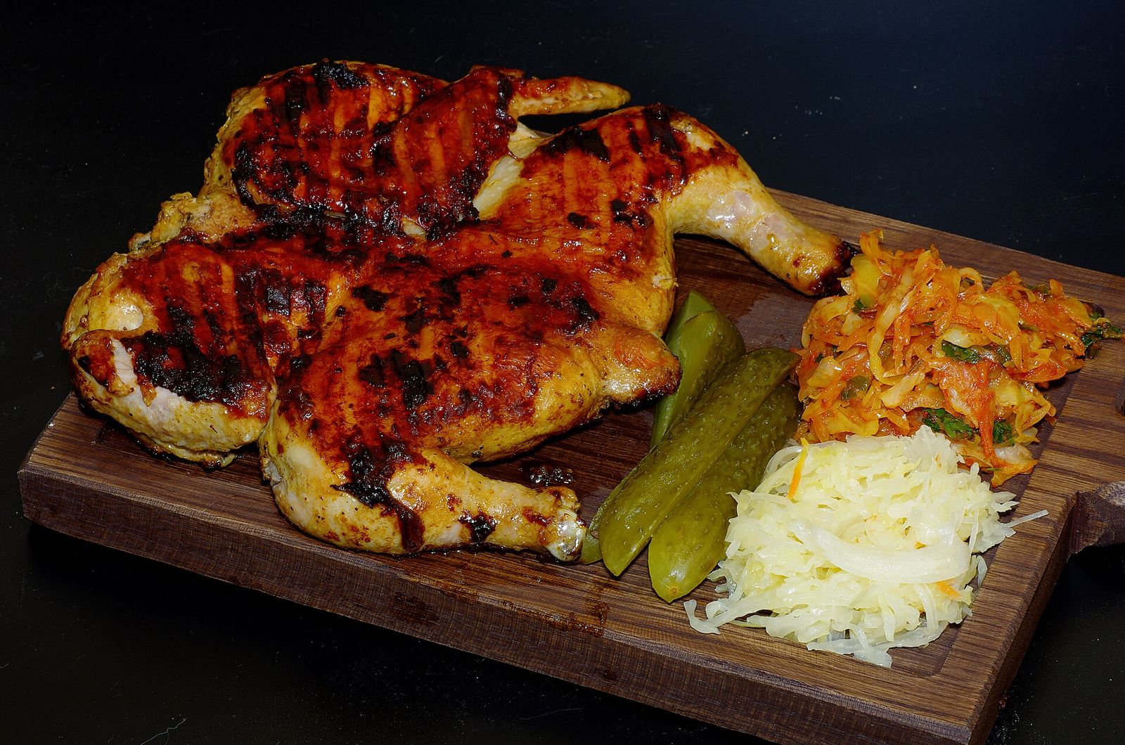 Chicken tabaka, a whole chicken pressed flat and fried until browned, on a wooden board with pickles and shredded cabbage