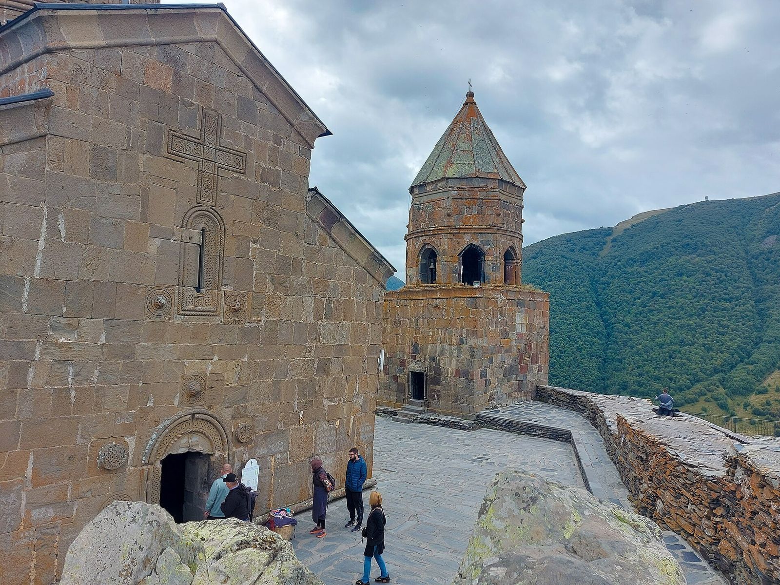 The carved stone wall of Gergeti Trinity Church and its separate bell tower, with visitors in the courtyard