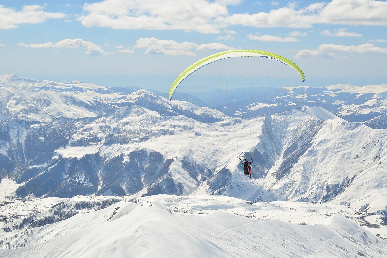 A paraglider with a yellow wing flying over snow-covered peaks above Gudauri