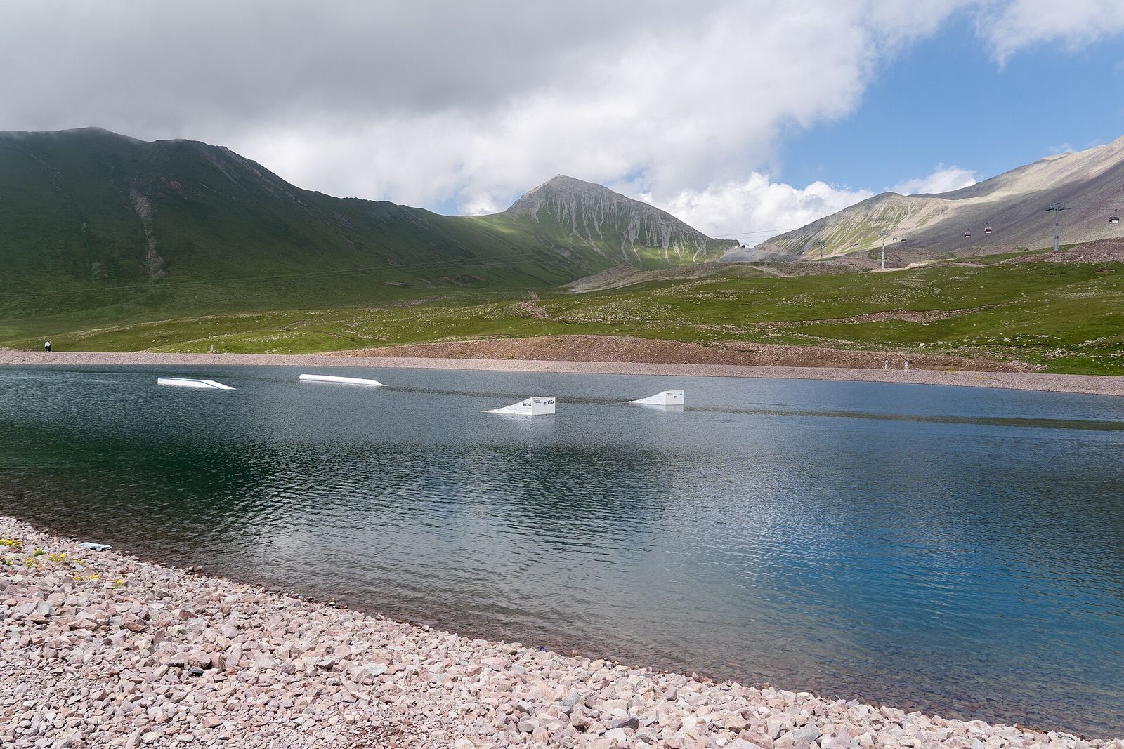 White Rabbit Wakeboard Park in Gudauri: white ramps on a blue mountain lake below green peaks, with a gondola line on the right