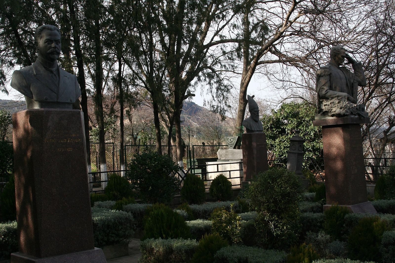 Bronze busts and a half-length uniformed figure on red granite plinths among clipped shrubs and trees at the pantheon of Azerbaijani figures in Tbilisi's National Botanical Garden