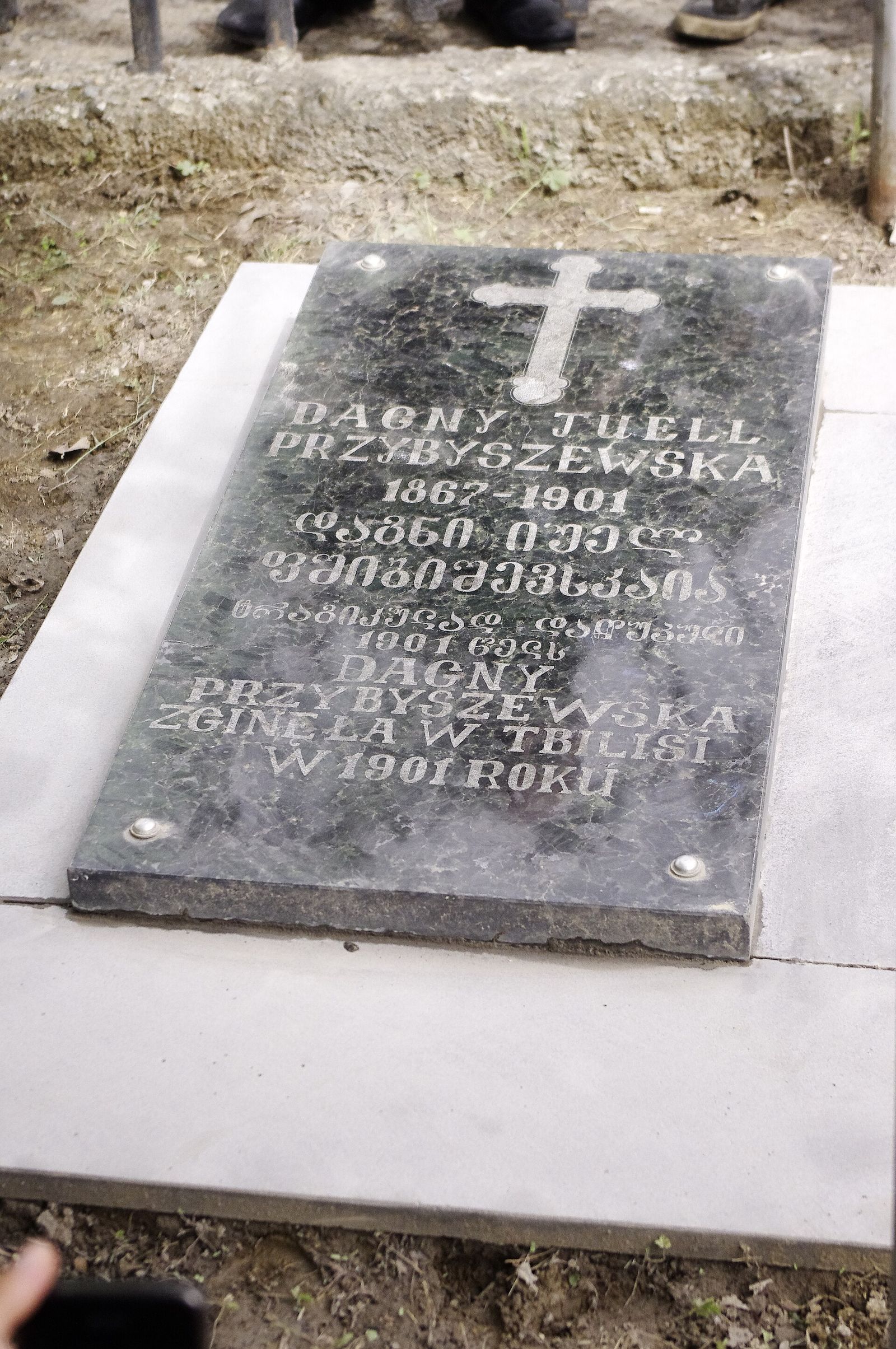 Dagny Juel's dark stone grave slab at Kukia Cemetery, a carved cross above her name in Latin and Georgian letters and the dates 1867 to 1901, with a Polish line saying she died in Tbilisi in 1901