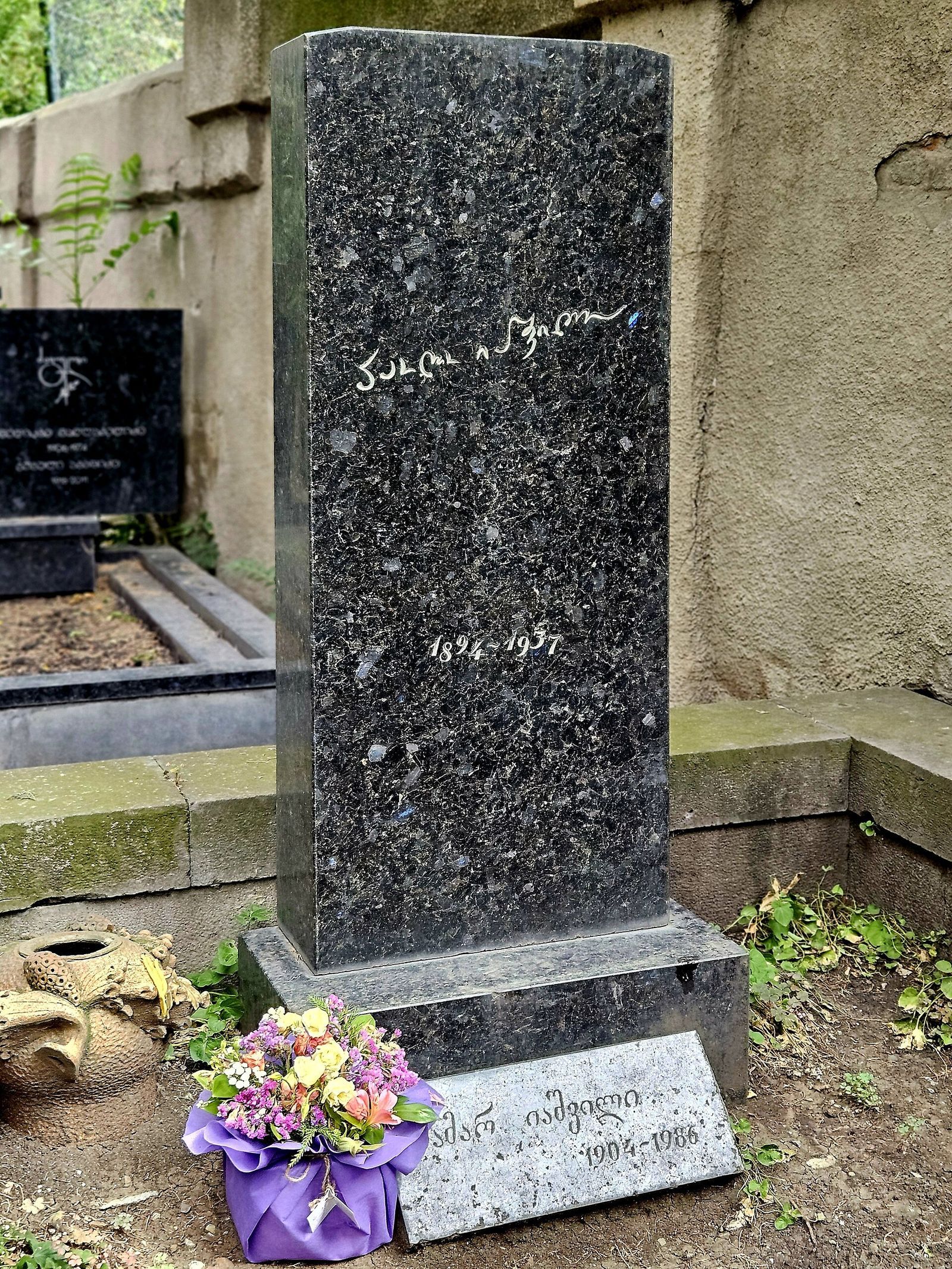 Paolo Iashvili's tall dark granite gravestone at the Didube Pantheon, his name in white handwritten Georgian script above the dates 1894 to 1937, with flowers at its base
