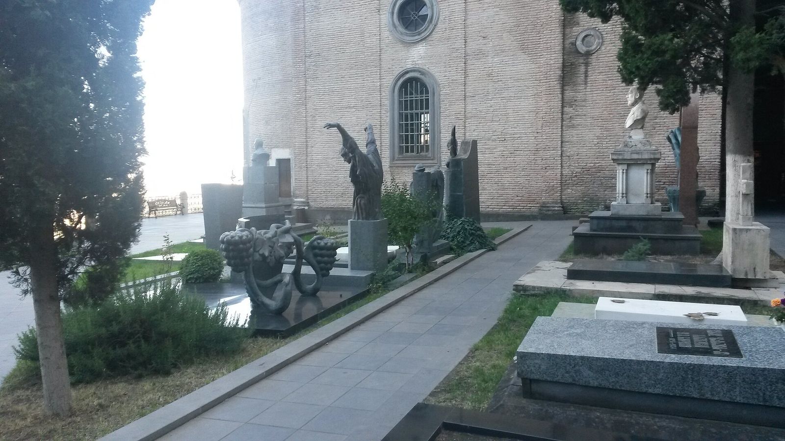 Sculpted gravestones, busts and polished slabs along a paved path beside the brick wall of Mamadaviti Church at the Mtatsminda Pantheon