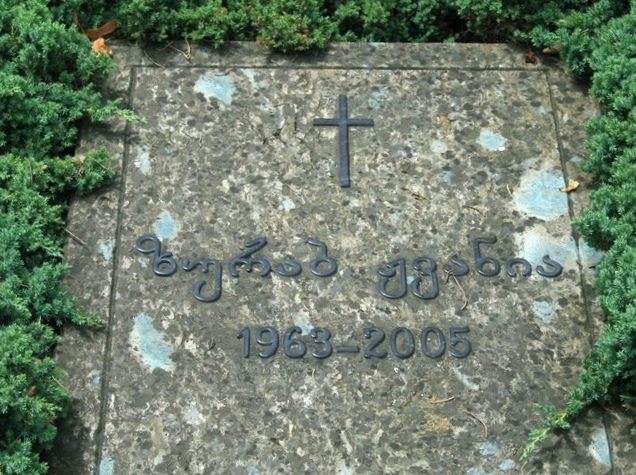 Zurab Zhvania's flat grave slab at the Didube Pantheon, a metal cross above his name in Georgian and the dates 1963 to 2005, framed by low juniper