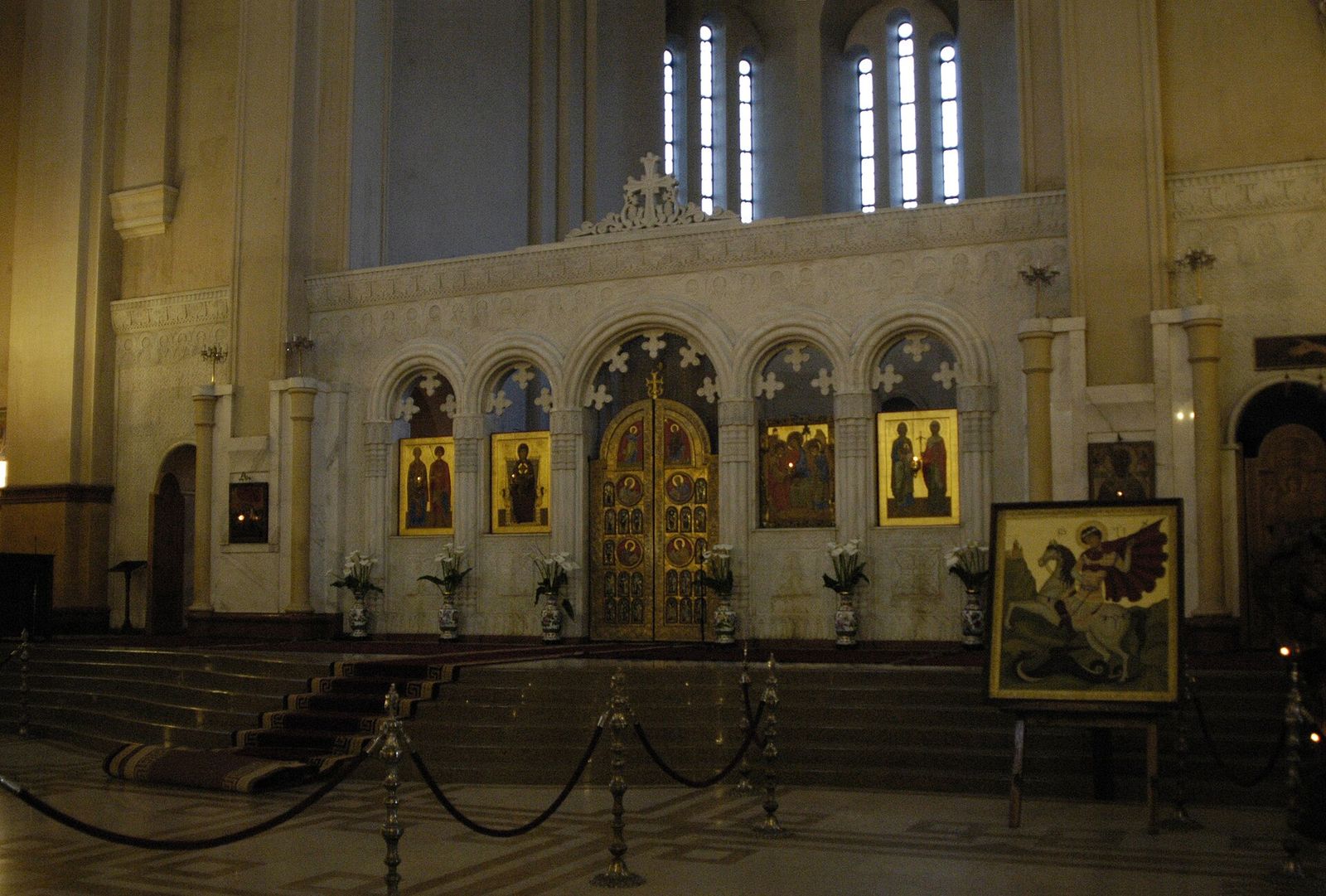 The pale stone iconostasis inside Sameba Cathedral, with golden royal doors and painted icons, and an icon of St George in the foreground