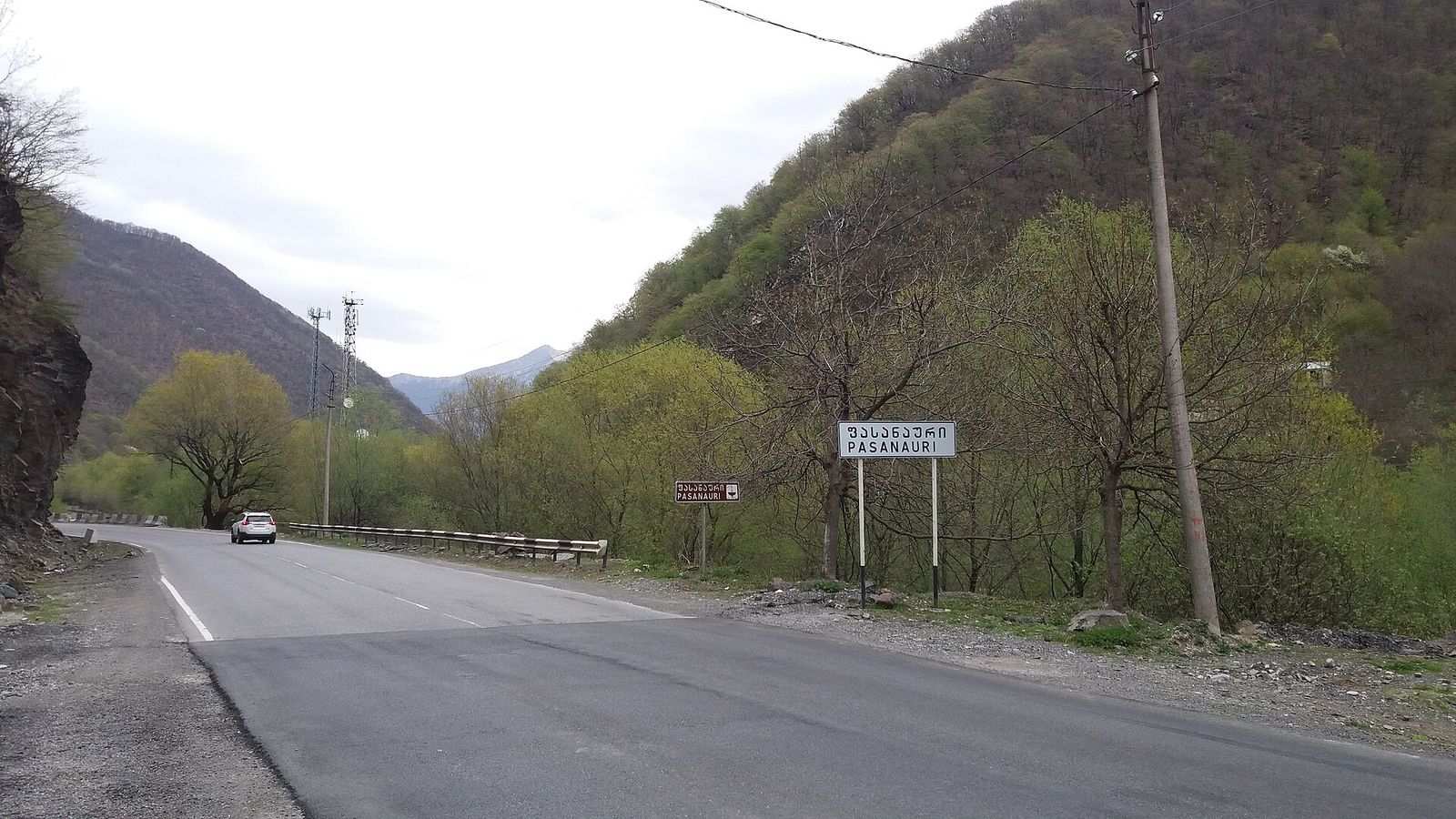 The Military Highway entering Pasanauri, with the town sign in Georgian and English beside the road and wooded hills behind