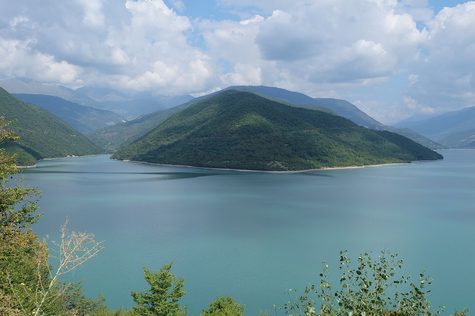 The turquoise Zhinvali Reservoir seen from above, with a forested hill and mountains behind