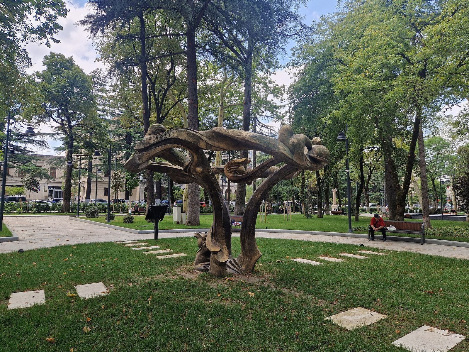 Lawns, paths and tall old trees in Kutaisi's central park, with a twisting memorial sculpture on the grass