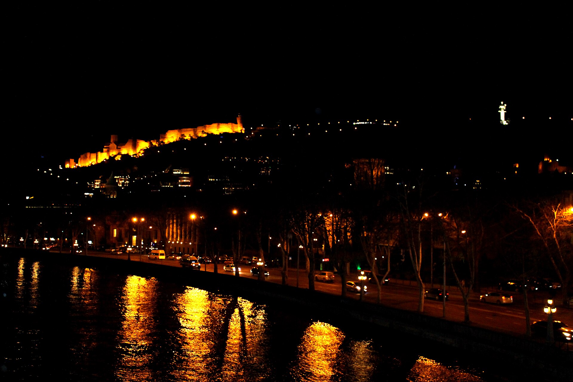 Narikala's floodlit walls and the lit Mother of Georgia on the ridge at night, seen from the Bridge of Peace across the Kura