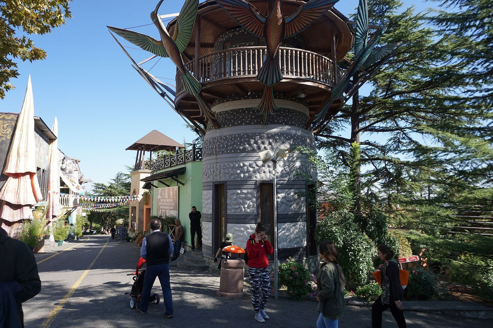Tower with giant carved birds on its balcony beside a paved walkway under cedar trees in Mtatsminda Park