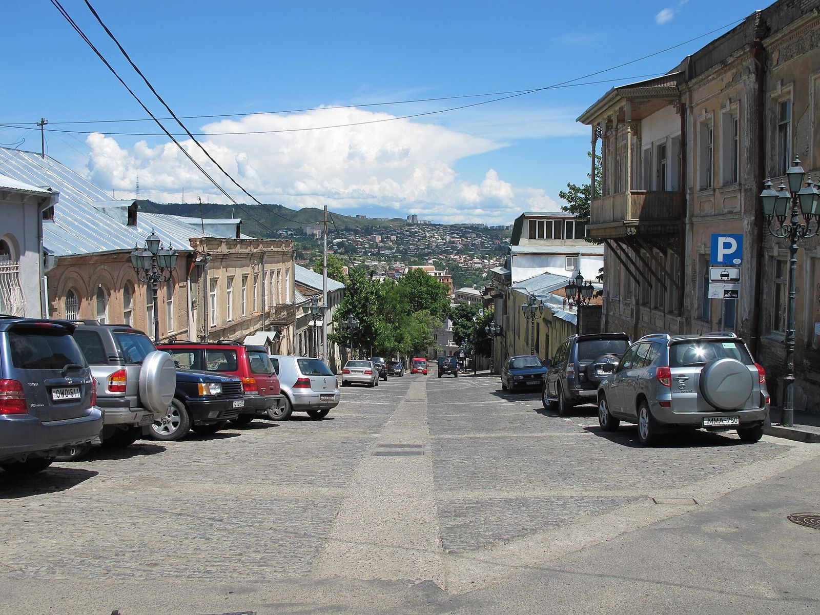Cobbled Mtatsminda Street sloping downhill between old houses with wooden balconies and iron street lamps, with the hills of the city beyond