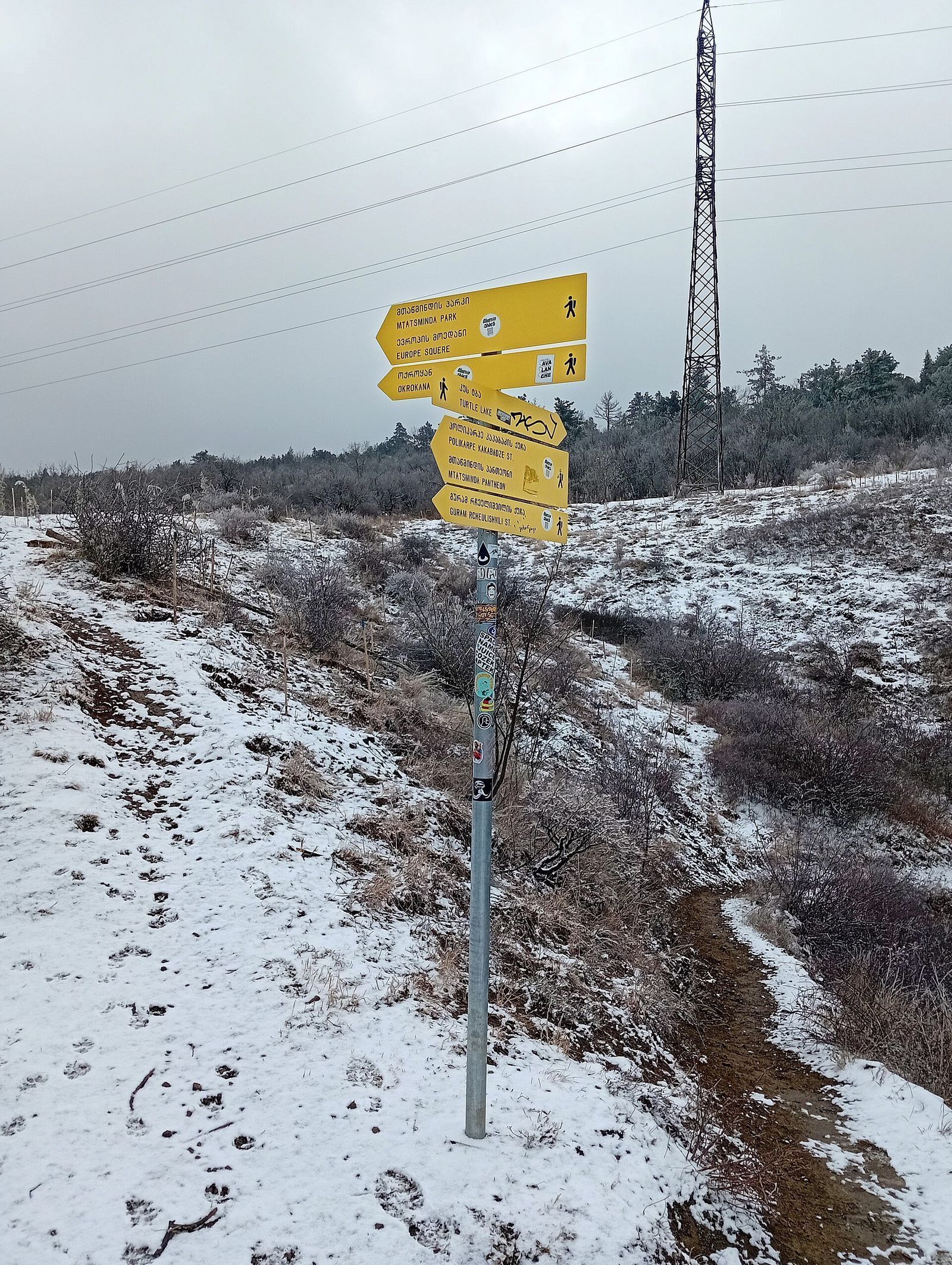 A yellow fingerpost on the Turtle Lake to Mtatsminda Park ridge trail, with signs for Mtatsminda Park and Turtle Lake, after a winter snowfall