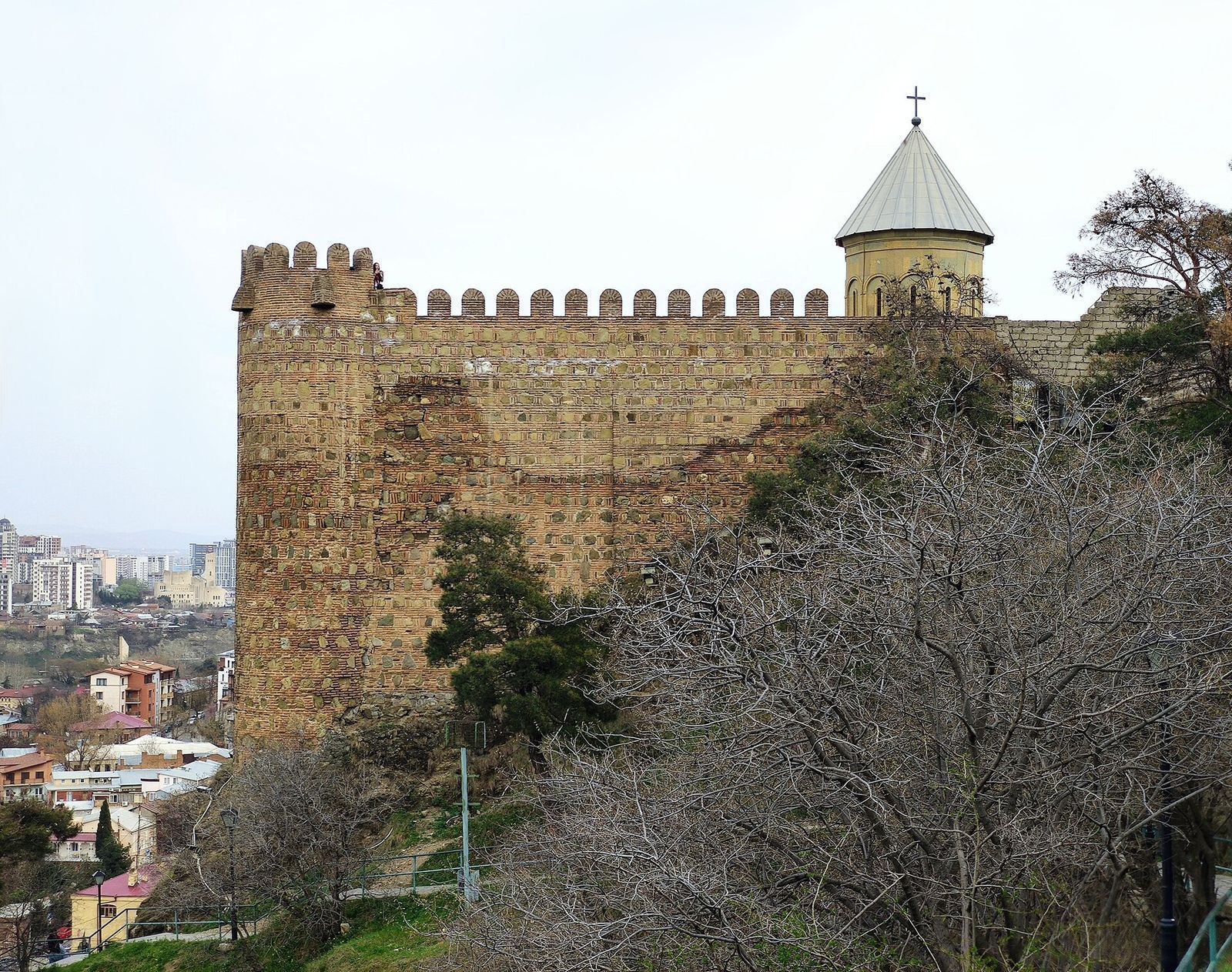A crenellated stretch of Narikala's lower walls with a round corner tower and the dome of St Nicholas Church behind