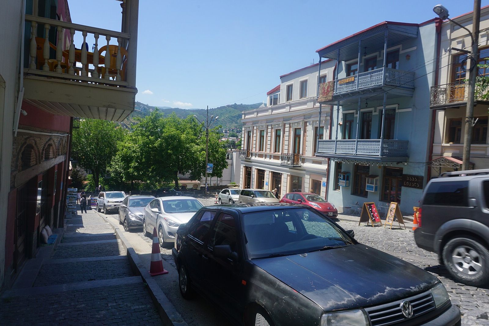A cobbled street in Old Tbilisi lined with parked cars and houses with blue wooden balconies, green hills behind