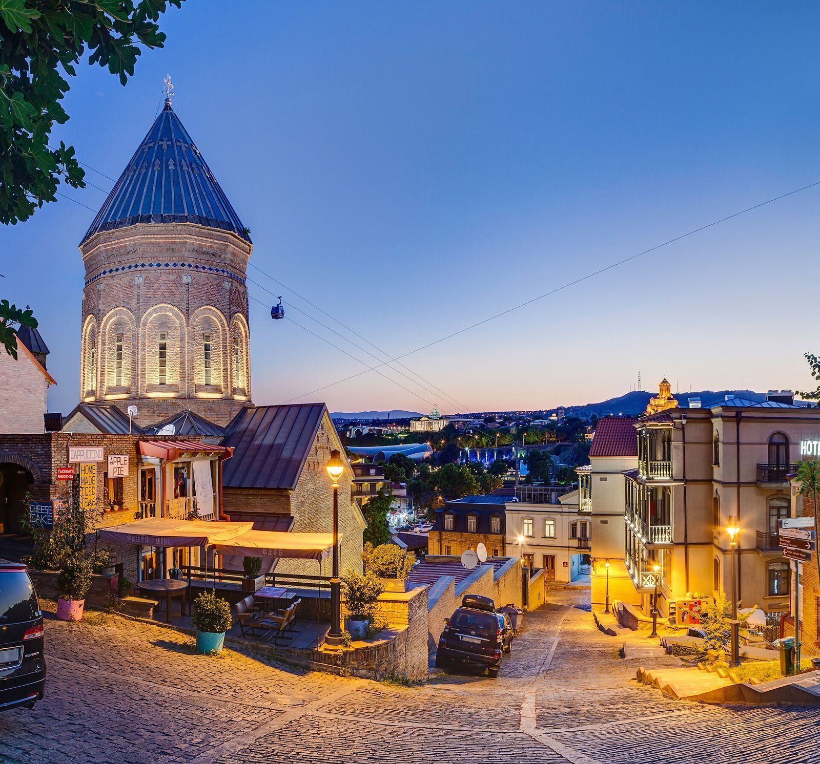 The cone dome of the Armenian church of Saint George above a cobbled lane in Old Tbilisi at dusk, with the awnings and terrace of Carpe Diem Café beside it