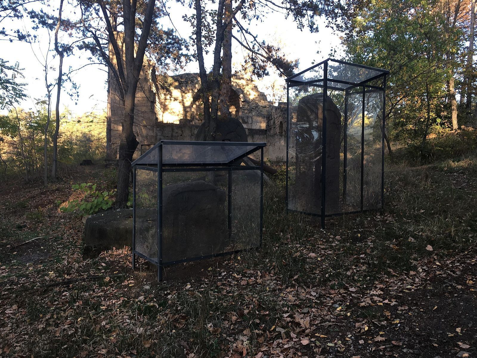 Carved stone stelae in glass cases among the trees at the Open Air Museum of Ethnography, with old stone walls behind