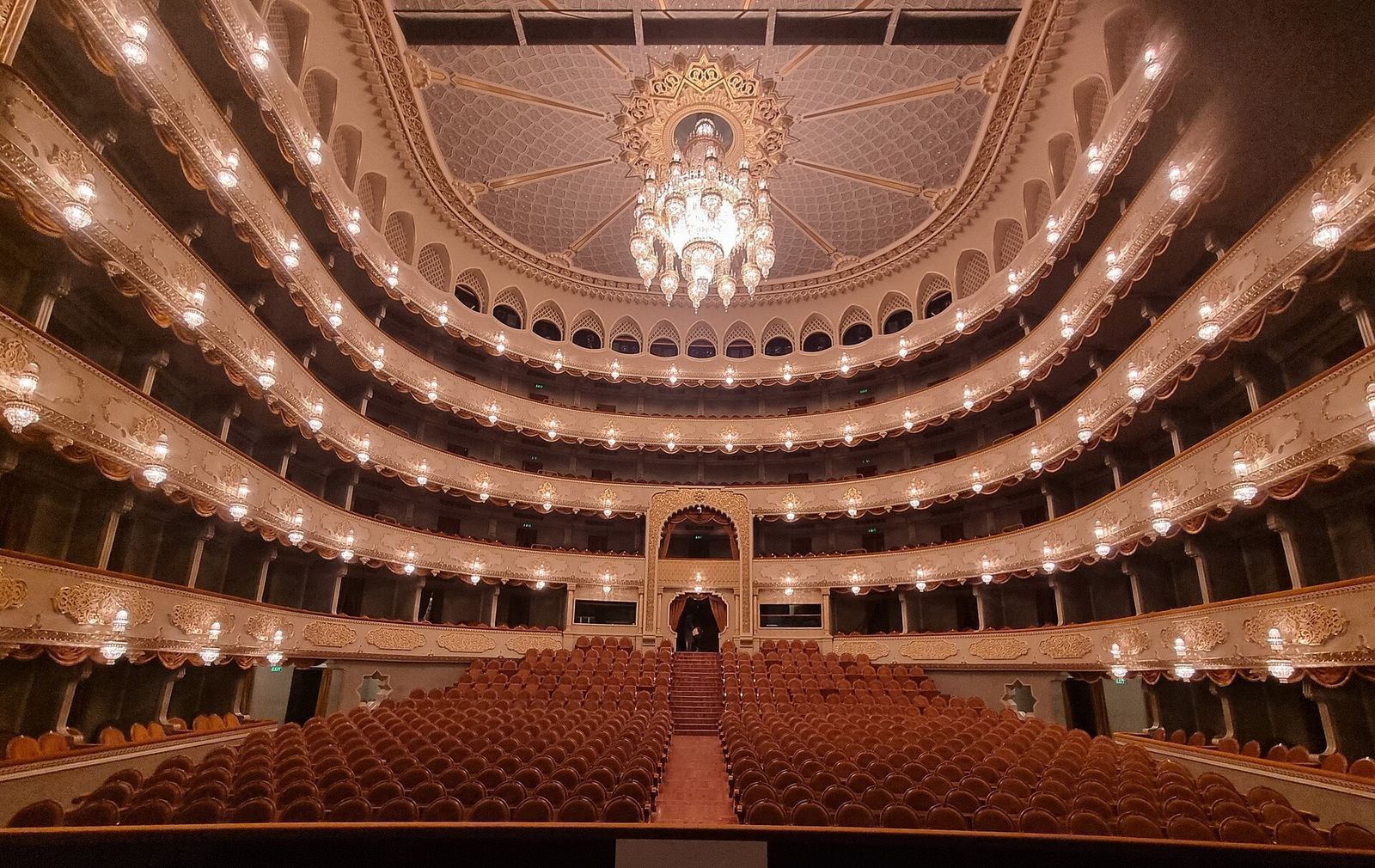 The auditorium of the Tbilisi Opera and Ballet Theatre seen from the stage, with red velvet seats, curved gilded balconies and the chandelier under the painted ceiling