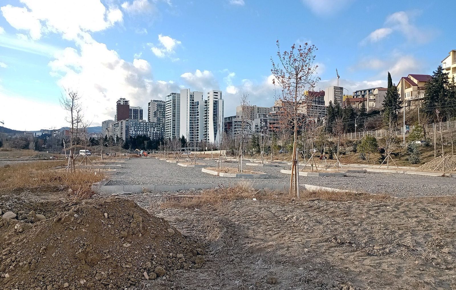 Open ground, new paths and young trees on the former Hippodrome in Saburtalo, with apartment towers behind