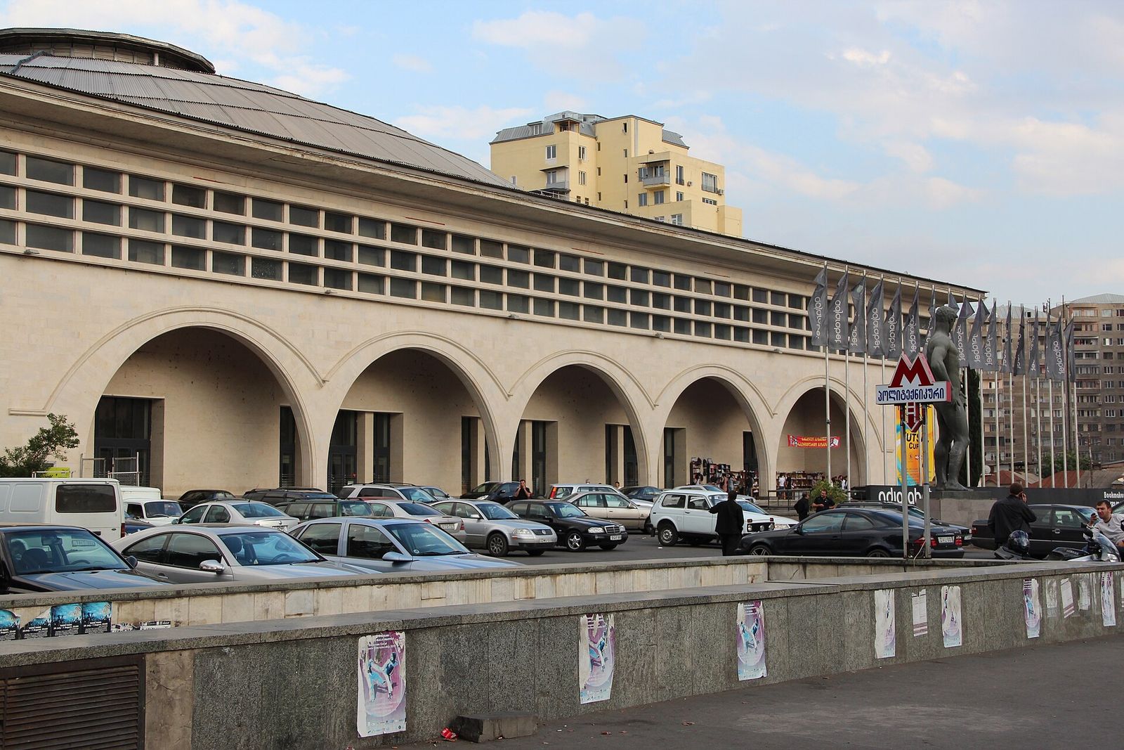 The arched facade and low dome of the Tbilisi Sports Palace in Saburtalo, with cars parked in front and a metro sign by the entrance