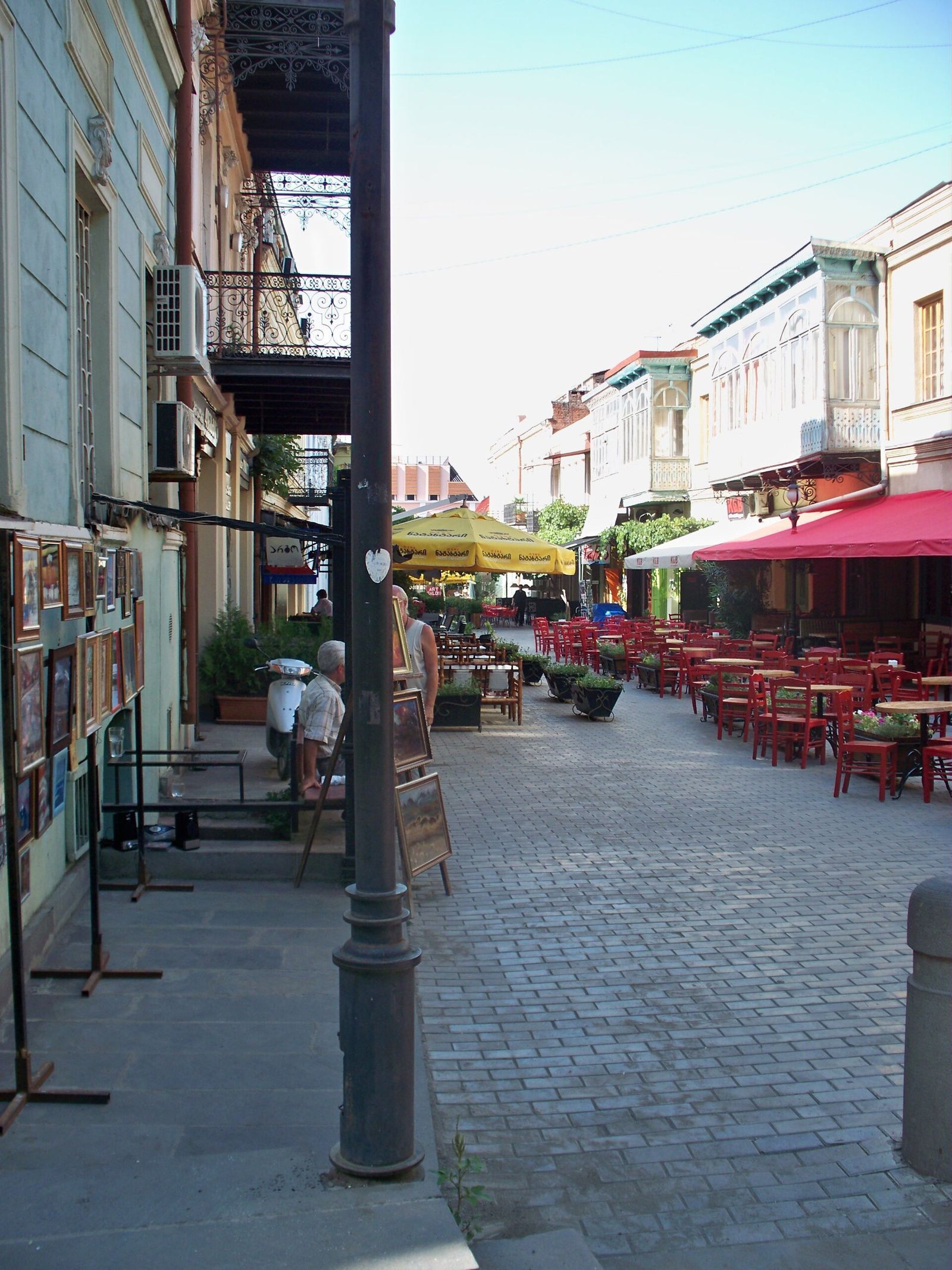 Paintings for sale on stands and walls outside an art shop on Erekle II Street, with cafe terraces further down the lane