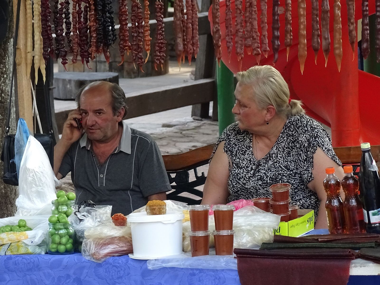 A couple at a street stall in Sighnaghi, with strings of churchkhela hanging behind a table of jars, bottles and green plums