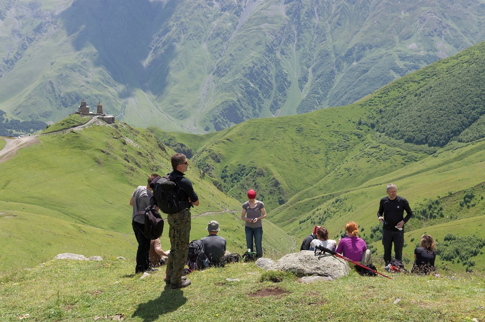 A group of hikers resting on a grassy ridge above Kazbegi, with Gergeti Trinity Church on the hill behind them