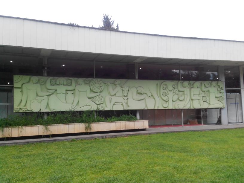 A long pale-green Soviet relief of farm workers, cattle and grapes along the front of the gallery pavilion at Expo Georgia, above a lawn