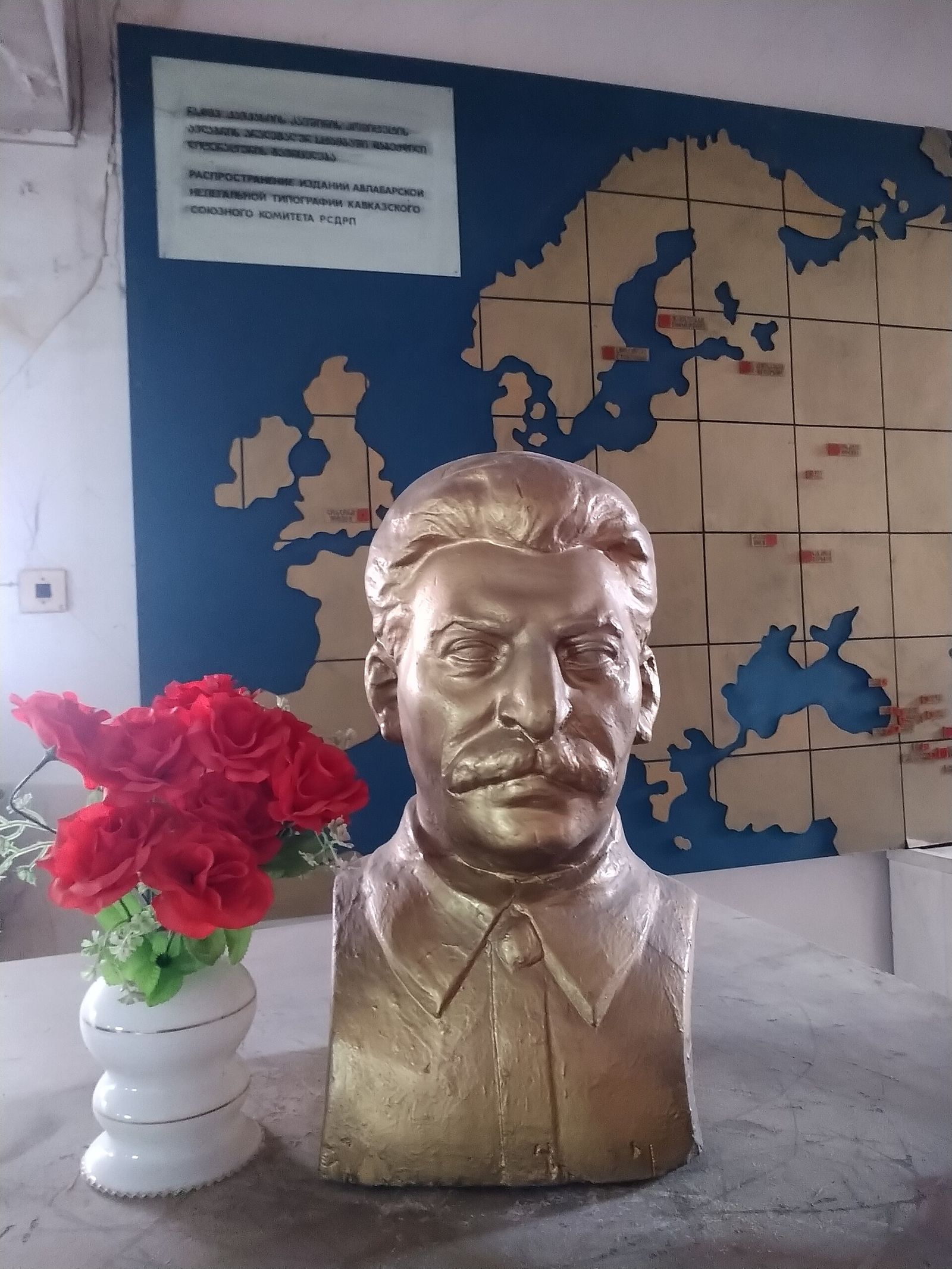 A gold bust of Stalin beside a vase of red roses, in front of the tiled map of Europe in the Underground Printing House Museum