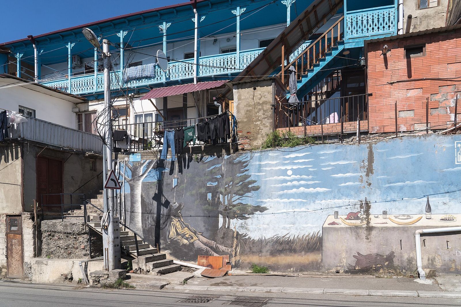A painted landscape mural on a street wall under blue wooden balconies and drying laundry on Mikheil Bukhaidze Street in Tbilisi