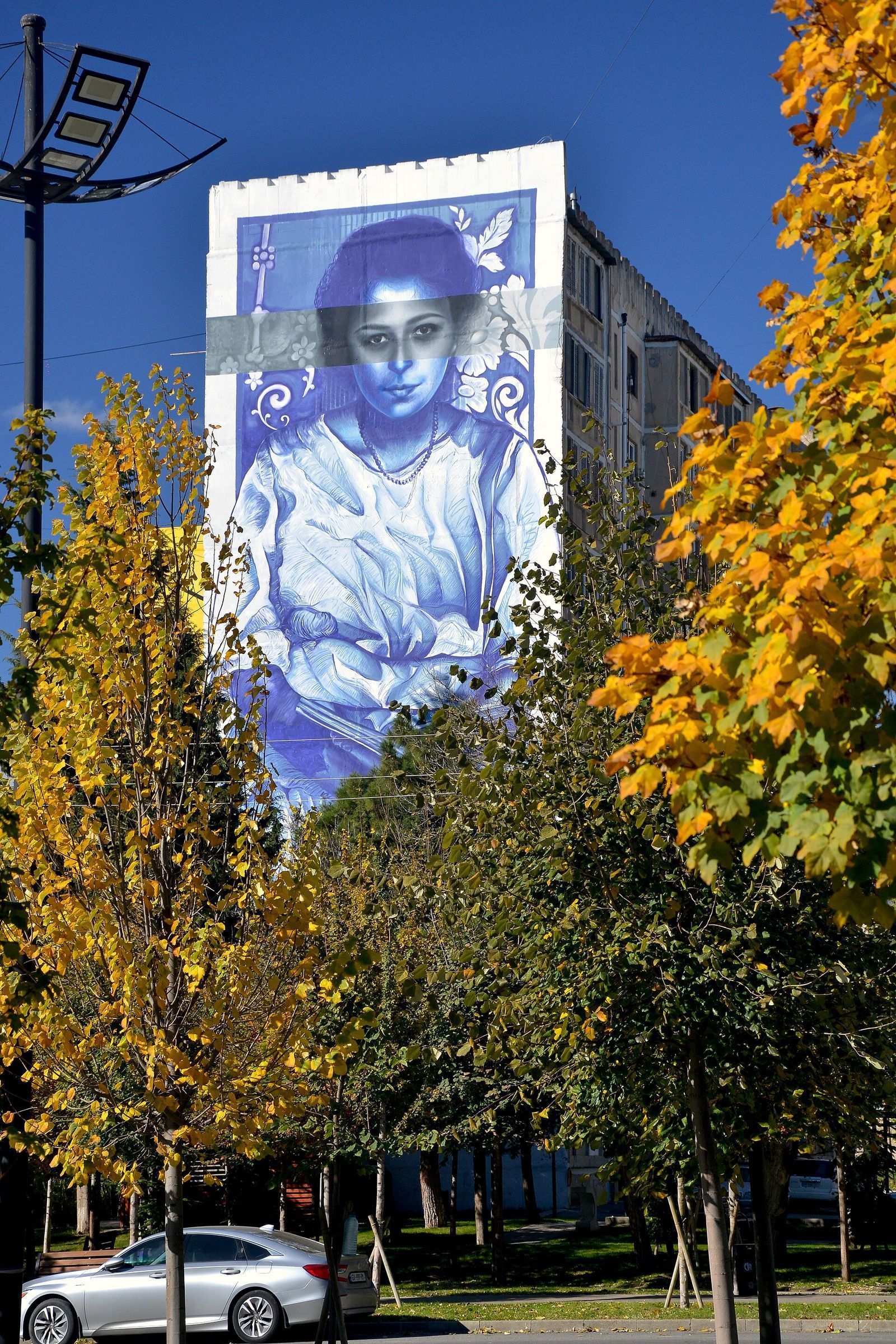A blue mural of Maro Makashvili covering the end wall of a Soviet apartment block in Varketili, Tbilisi, behind autumn trees