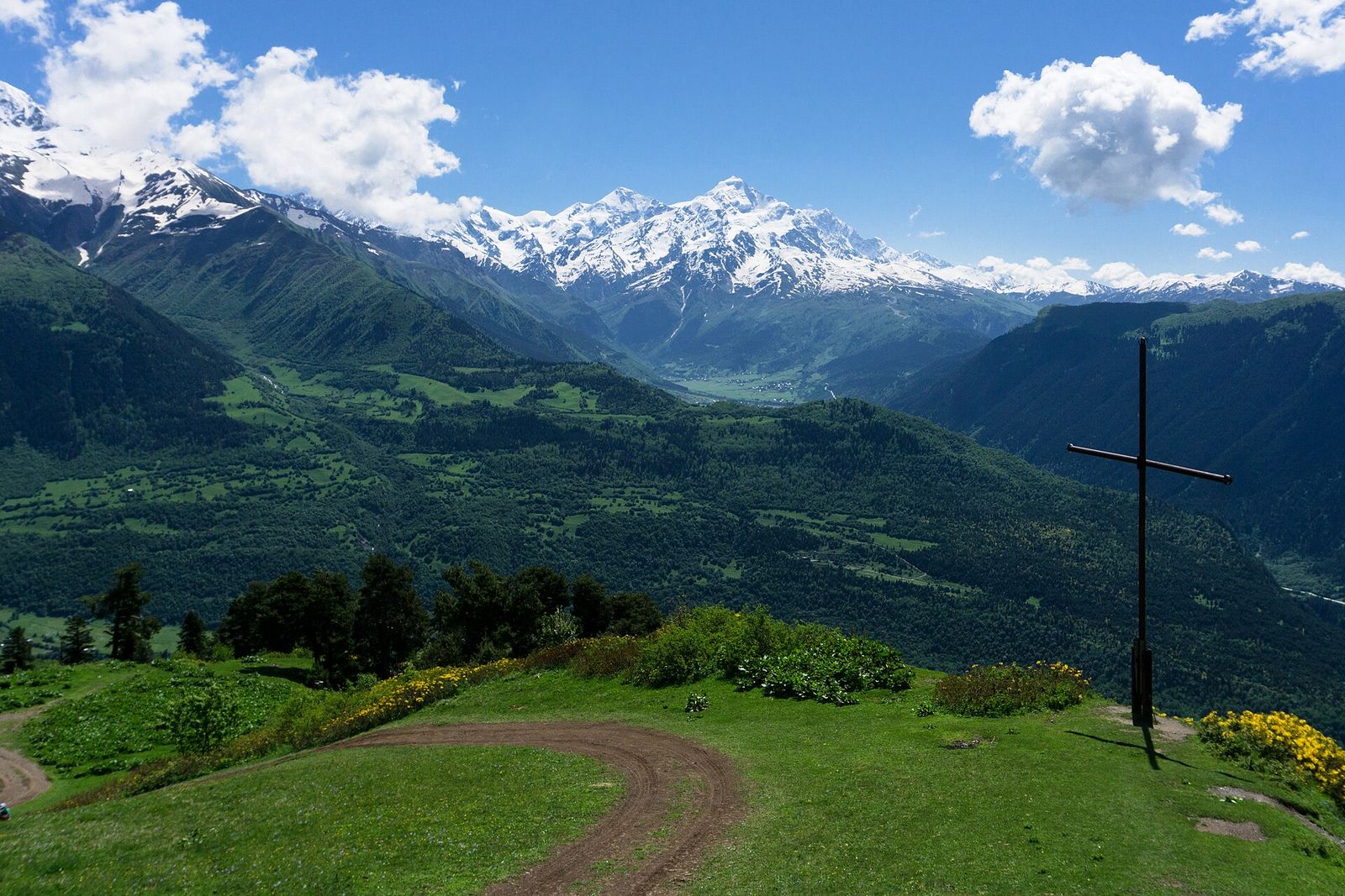 The metal cross on the grassy ridge above Mestia, with the jeep track, yellow azaleas and snowy Caucasus peaks across the valley