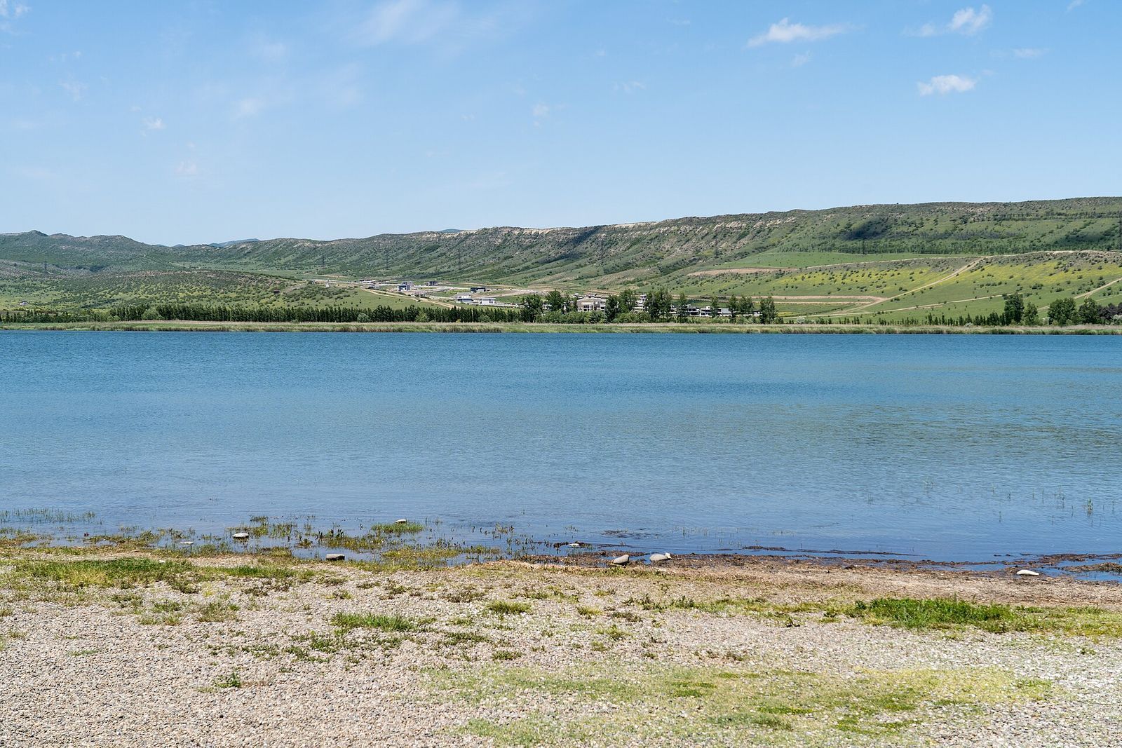 The pebbly shore and blue water of Lisi Lake in late spring, with green hills on the far side