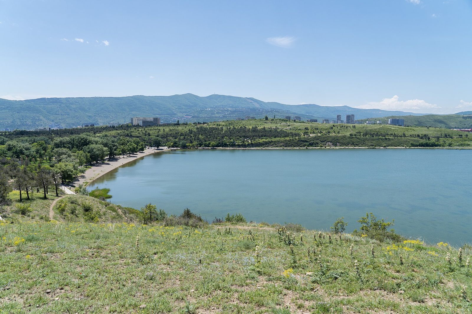 Lisi Lake in summer, with a curved beach and lakeside path on the left and green hills and apartment blocks behind
