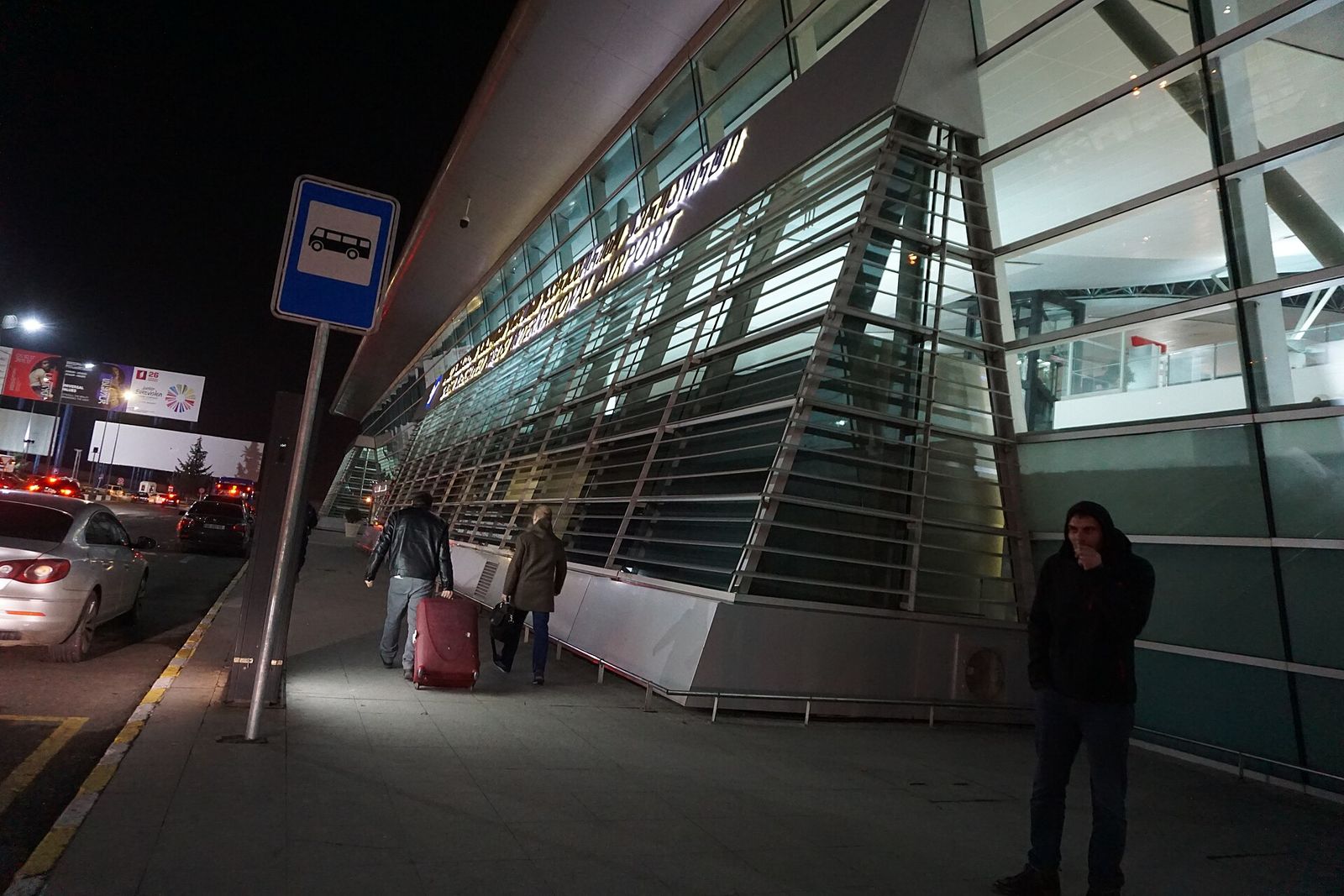 The bus stop sign outside Tbilisi International Airport at night, with travelers wheeling suitcases past the lit terminal