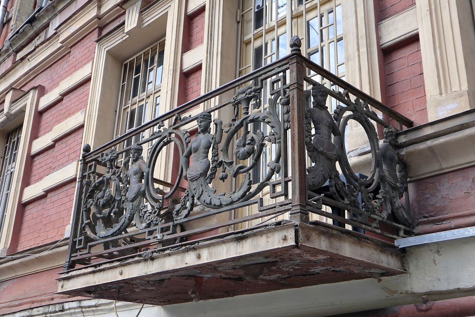 A cast-iron balcony with sculpted female figures and scrolls on a brick house at Jerusalem Square 6, Tbilisi