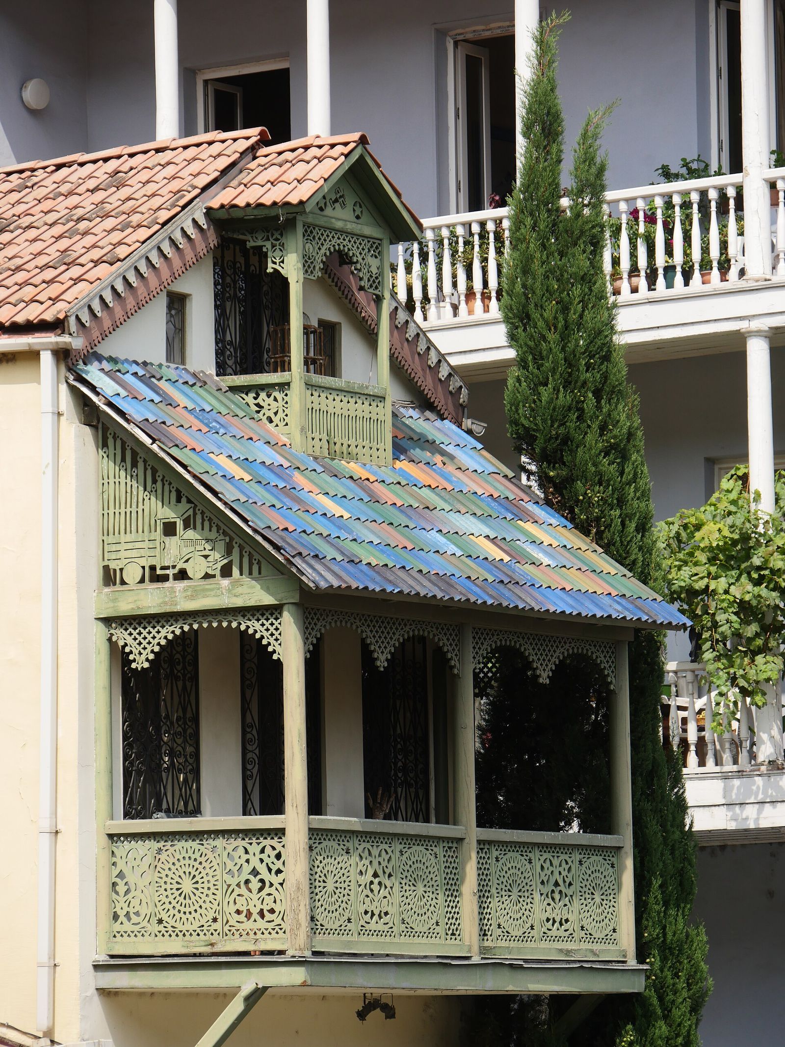 The green carved wooden balcony on Abano Street, Tbilisi, with a truck and a biplane cut into the fretwork under a multicolored roof