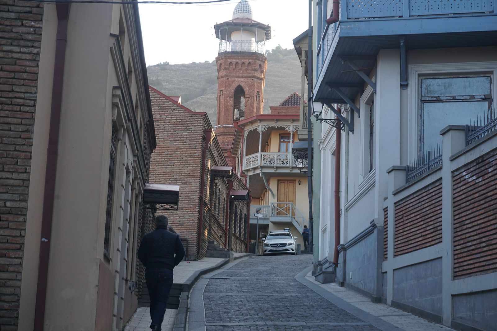 Botanikuri Street in Tbilisi climbing toward the brick minaret of the Jumah Mosque, with a carved wooden balcony at the top of the lane