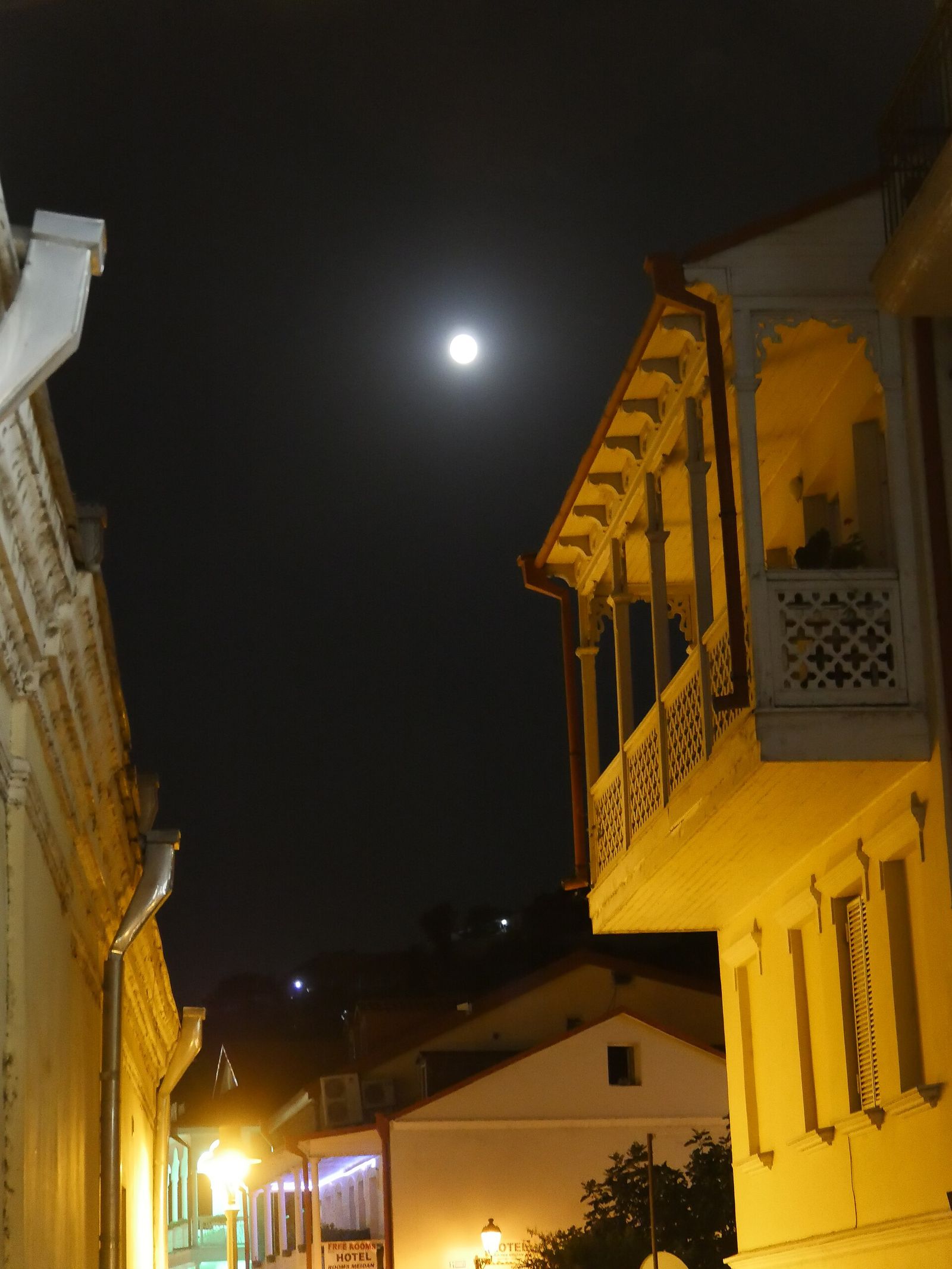 A carved wooden balcony lit up at night on Samghebro Street, Tbilisi, under a full moon