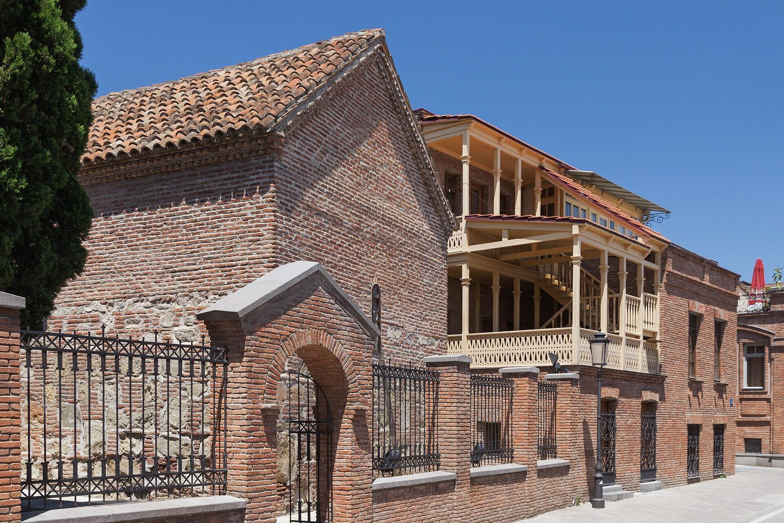A restored wooden balcony with carved railings beside the brick Kari church on Ioane Shavteli Street, Tbilisi
