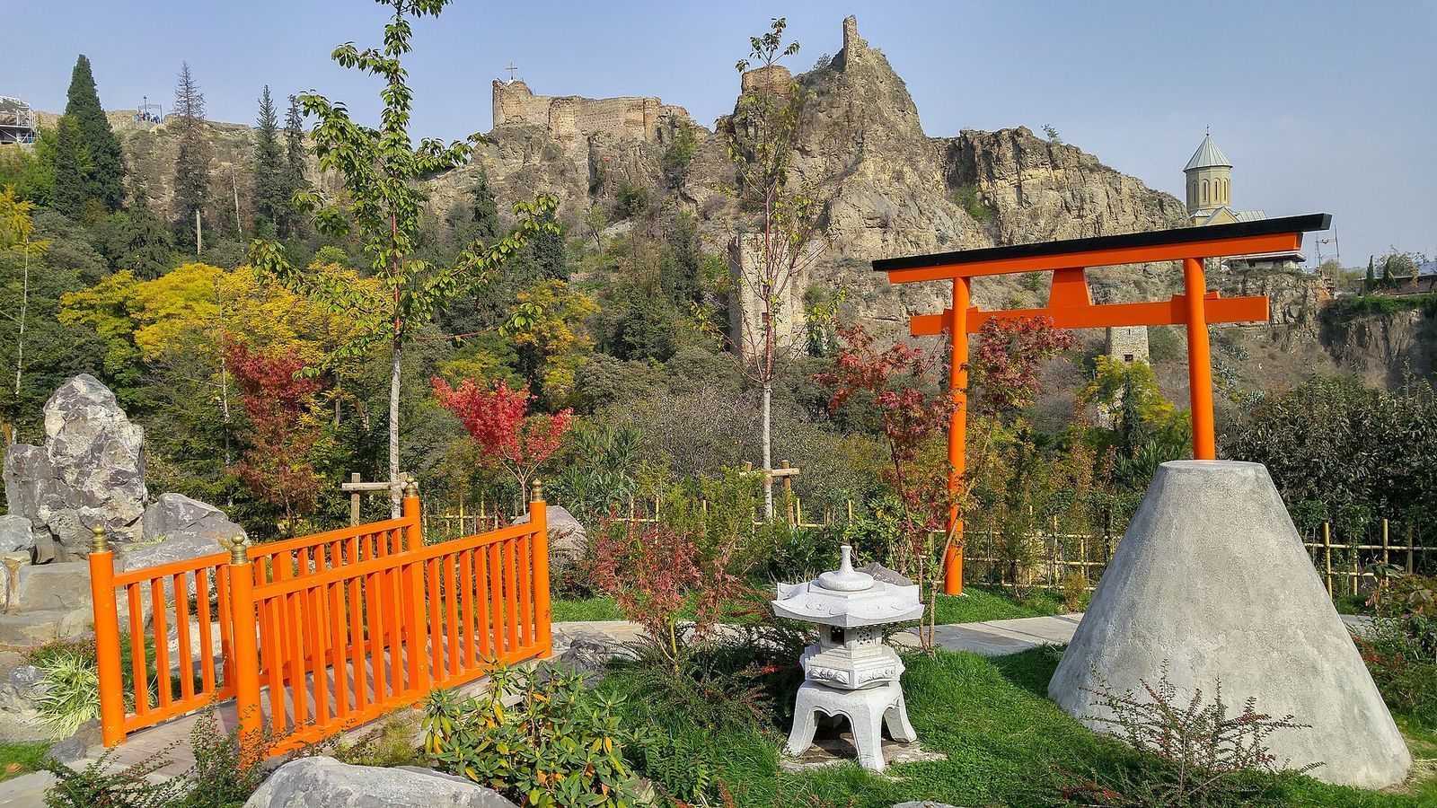 The Japanese garden in the Tbilisi Botanical Garden, with an orange torii gate, a stone lantern and a footbridge below the walls of Narikala