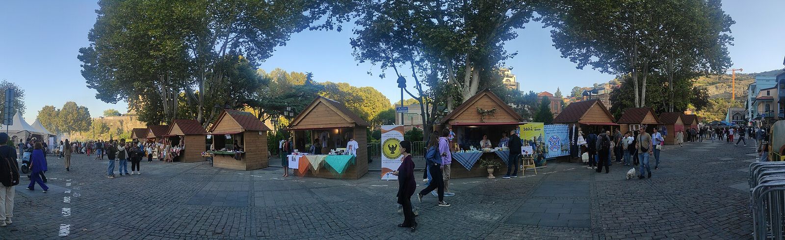 Wooden festival stalls under tall plane trees on a cobbled square in Abanotubani during Tbilisoba 2024