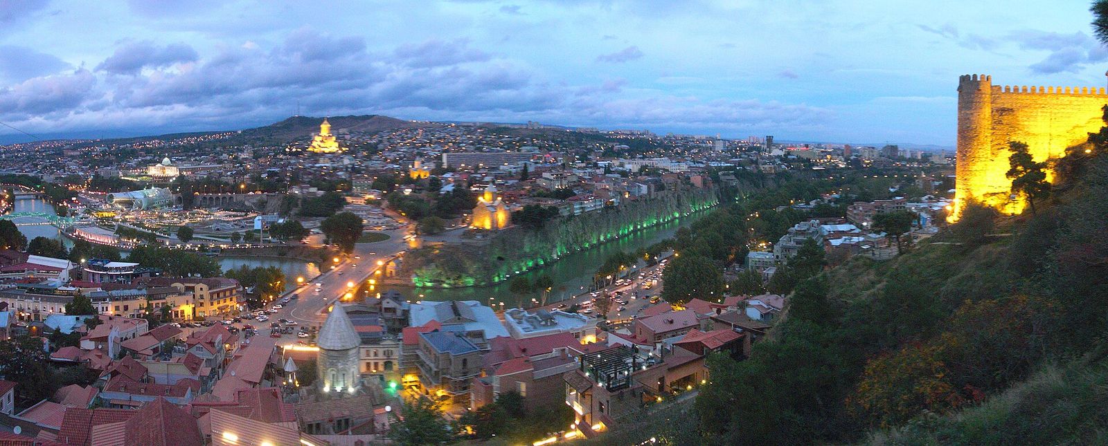 Dusk view from the Narikala ridge over the Old Town rooftops, the river, Rike Park and the lit-up Holy Trinity Cathedral, with the floodlit fortress wall on the right