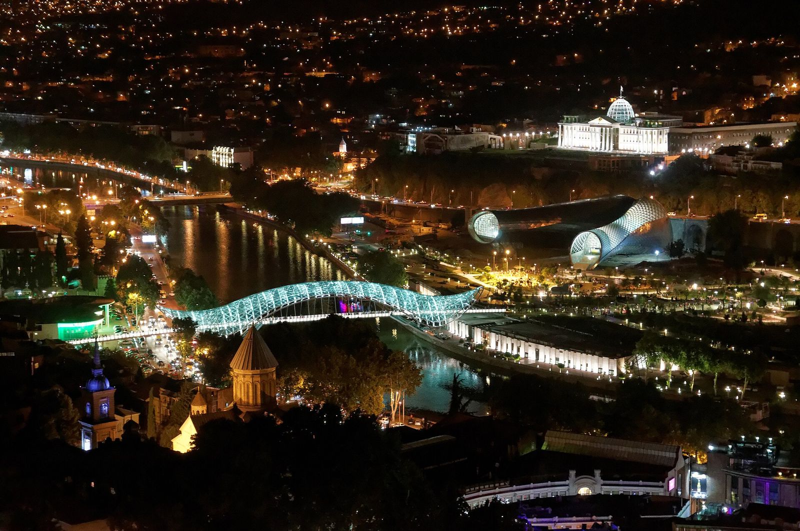The Bridge of Peace lit up over the Mtkvari River at night, with Rike Park beyond it