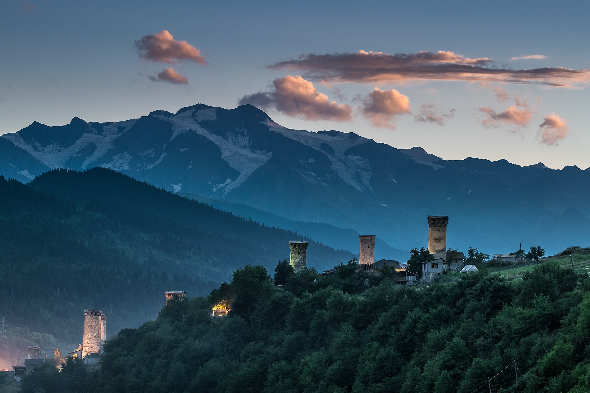 Mestia's medieval stone defense towers at dusk, with forested slopes and snow-streaked Caucasus peaks behind