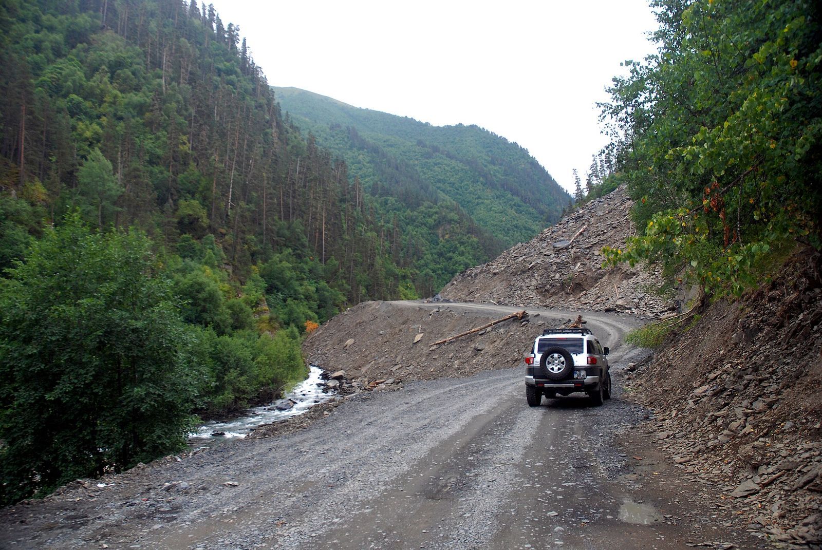 A white 4x4 on the unsealed Tusheti road in a forested gorge, with landslide debris on the slope beside it